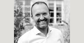 Black-and-white portrait of a smiling bearded man in a light shirt, standing outdoors before a blurred building