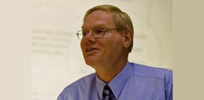 Middle‑aged man with glasses in a light blue shirt and striped tie, smiling and looking sideways indoors