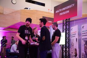 People talk at an Aalto University pavilion stand indoors with pink lighting and banners.