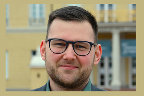 Smiling man with dark hair, beard and black glasses outdoors in front of a yellow building