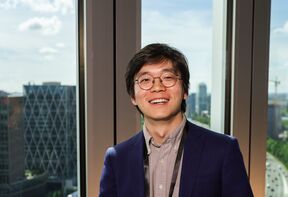 Smiling person with glasses in a blue blazer stands indoors by tall windows overlooking a city skyline.