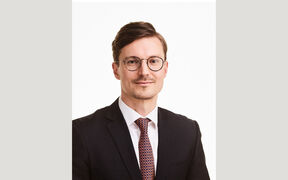 Portrait of a man in a dark suit, white shirt and patterned tie, wearing round glasses, against a light background