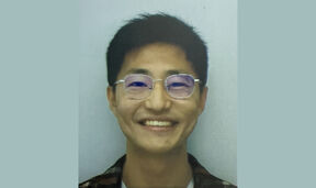 Smiling young man with short dark hair and round glasses, facing the camera against a pale background