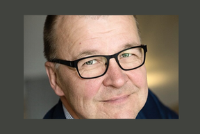Middle‑aged man with black glasses smiling softly, close‑up indoors with neutral background