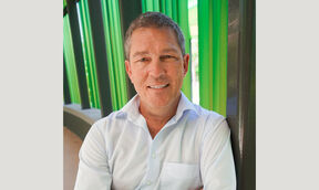 Smiling middle‑aged man in a white shirt, seated by tall green and dark columns outdoors