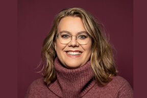 Smiling woman with glasses and wavy blond hair in a burgundy polo-neck jumper, against a matching plain background