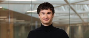 Smiling man in a black turtleneck stands indoors before glass walls and bright ceiling beams