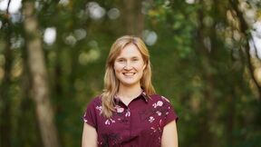 Smiling woman in a patterned burgundy shirt standing outdoors against blurred trees