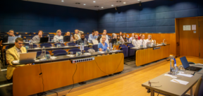 Small audience in a modern lecture hall, seated with laptops and water bottles, facing a table at the front