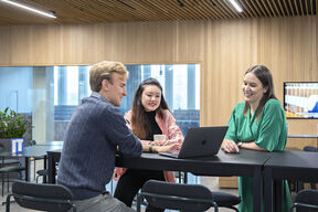 Three colleagues sit at a tall office table, chatting and looking at a laptop together