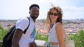 Two smiling tourists with backpacks pose by a city viewpoint, rooftops and greenery behind them
