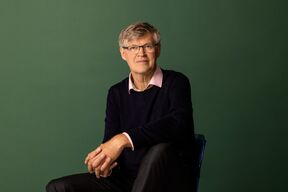 Middle‑aged man with glasses seated, wearing dark jumper and pink shirt, against plain green background.