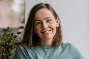 Smiling woman with straight brown hair in a green top, sitting indoors with plants in the background