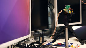 Three monitors on a desk. A camera on a tripod focuses on a circuit board.