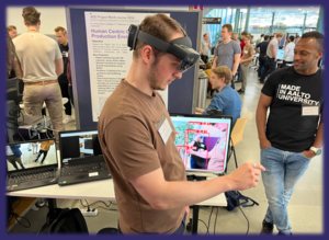 A man wearing a brown shirt uses virtual reality equipment in a crowded event. Another man wears a black t-shirt with white text.