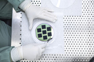 A person in protective gear handling a silicon wafer with black squares using tweezers in a cleanroom.
