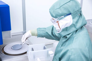 Scientist in green cleanroom suit handling a grey silicon wafer with tweezers at a lab machine