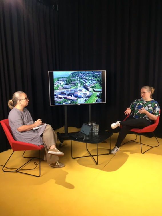 kaksi naista istuu studiossa punaisilla tuoleilla keskustelemassa, heidän välissään on näyttöruutuon 