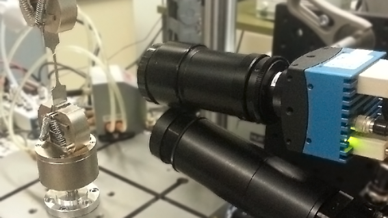 A metal sample clamped in a tension machine, being photographed by a high speed camera
