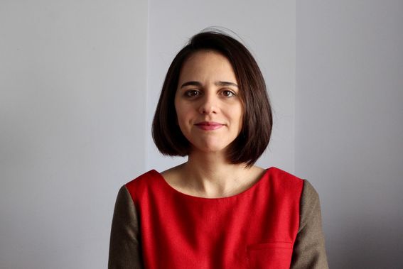 Tummatukkainen nainen katsoo suoraan kameraan, hänellä on punainen mekko yllään ja taustalla vaalea seinä