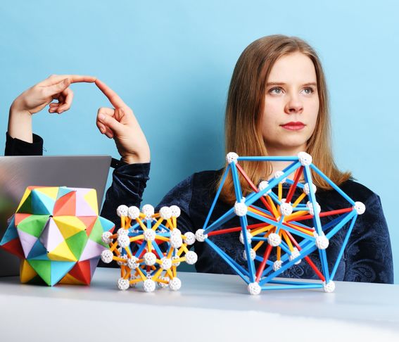 Nainen katsoo oikealle ja koskettaa kuvan vasemmalta tulevaa kättä. Etualalla värikkäitä geometrisiä kappaleita