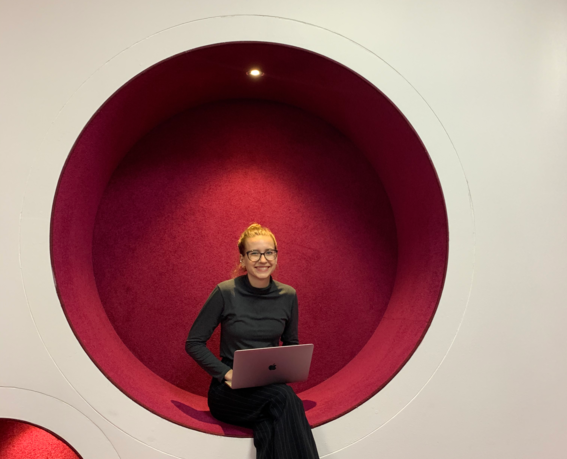 Svitlana Chaplinska sitting with her laptop and smiling towards the camera in a futuristic, round and red chair integrated to the wall at Aalto University campus.