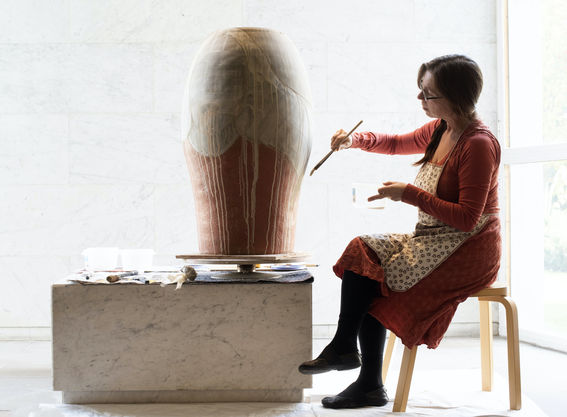 Professor Maarit Mäkelä sits on a chair and paints a ceramic pot with soil from Venice, in a brightly lit area