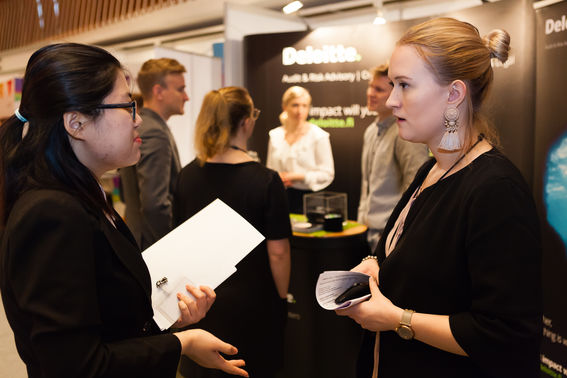 Two women holding papers and talking to eachother next to a group of people