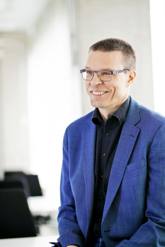 Keski-ikäinen mies hymyilee. Hänellä on sininen pikkutakki ja mustasankaiset silmälasit.