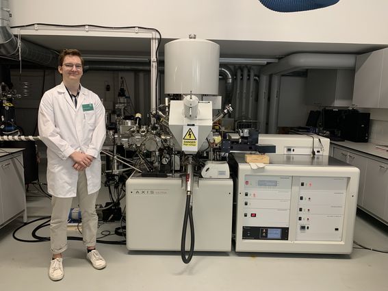 Color photo of a scientist wearing a white lab coat and glasses standing next to a large XPS device in a lab