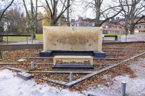 An outdoor artwork made of plywood, installed next to shopping centre A Bloc. It's autumn and there are brown leaves on the ground, while the grass on the background is still green.