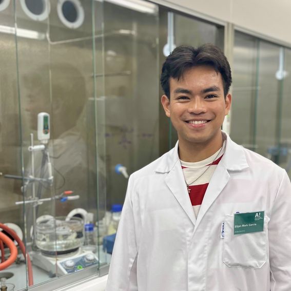 Color photo of a smiling young adult with short black hair wearing a lab coat and standing in front of experimental equipment in a lab
