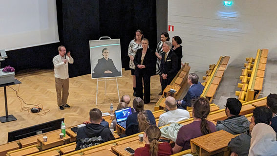 Professori Kaisa Nybergin muotokuvan paljastustilaisuus, auditoriossa on ihmisiä ja lavalla muotokuva ja Kaisa Nyberg.