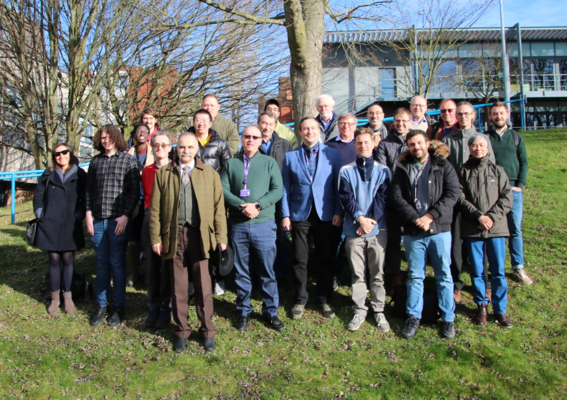 A group of 23 pose for a picture outside on a green hill with buildings in the background.