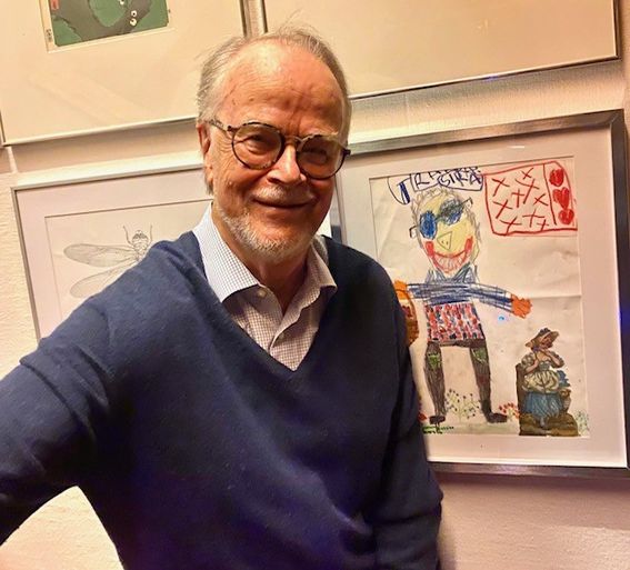 A man looks at the camera, a picture of him drawn by a child is framed on the wall behind him