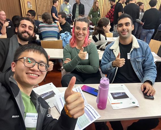 A group of people indoors, some sitting at desks with papers in front of them, giving thumbs up.