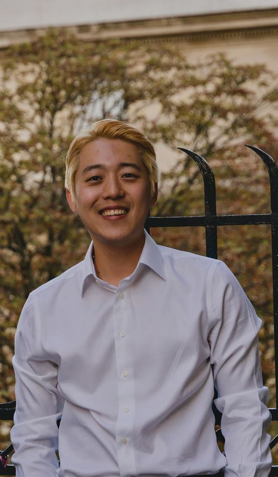 A person in a white shirt sits in front of a black fence with trees in the background.