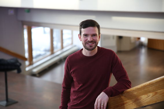 A person in a maroon jumper standing indoors, leaning on a wooden railing. Snow seen outside through the windows.
