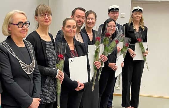 Group of people dressed in black holding certificates and roses. Some wearing academic caps indoors with grey background.