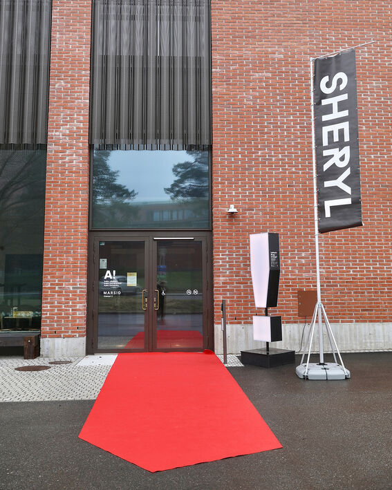 Entrance to a brick building with a red carpet, large windows, and a banner displaying 'Sheryl'.