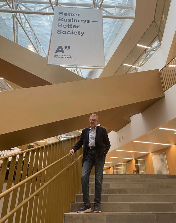 Matti Lehti standing at the School of Business stairway. Behind him, there's a Better Business - Better Society poster.