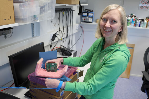 A person in a green hoodie holds a computer component in a workspace with boxes, a monitor, and various tools.