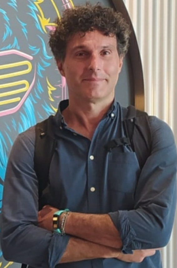 Man in a dark blue shirt with rolled-up sleeves, standing with arms crossed, colourful mural in background.