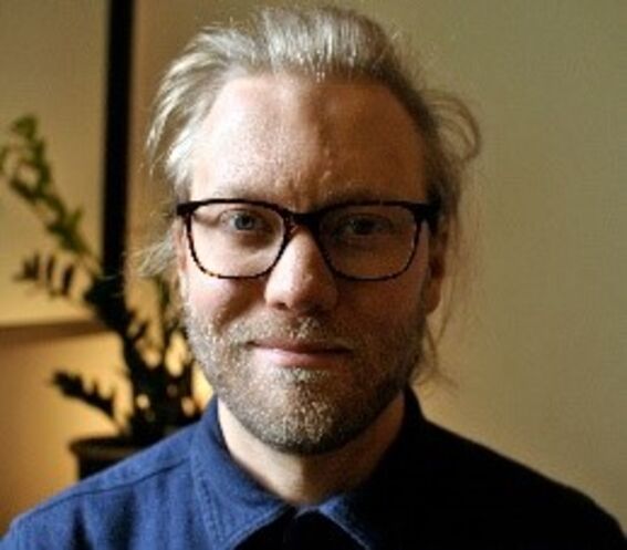 Person with light blond hair in a blue shirt, indoor setting with a plant in the background.