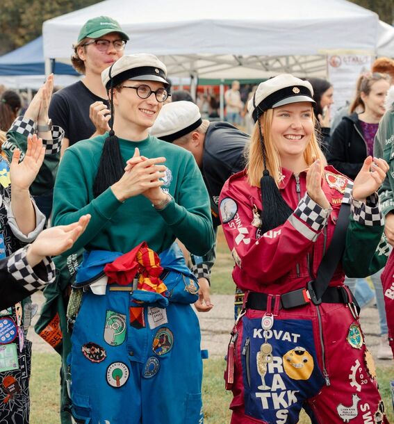 A group of university students dressed in colourful student overalls decorated with patches and accessories, clapping outdoors.