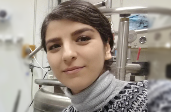 Person wearing a patterned knit sweater and grey turtleneck in a science laboratory with metal equipment in the background.