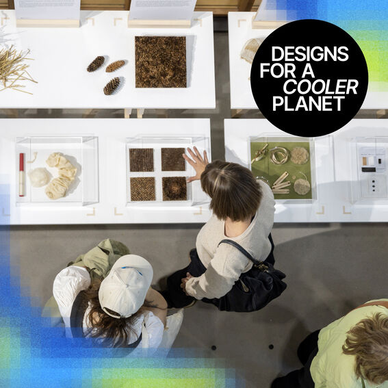 In this bird’s-eye view photo, three people are exploring the Designs for a Cooler Planet exhibition. They are examining materials and accompanying descriptions displayed on white tables. In the top right corner of the image, there is a black circle with the white text "Designs for a Cooler Planet" inside. In the bottom left corner, there is a section of a blue-green heat map.
