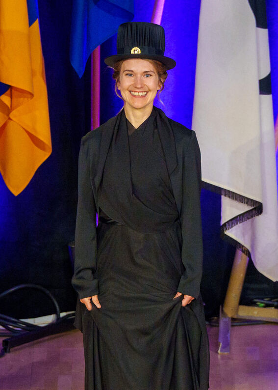 A person in a black academic gown and cap stands in front of coloured flags.