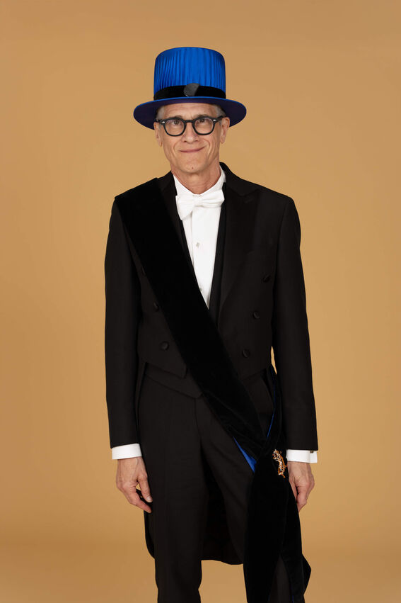 Henkilö mustassa juhlapuvussa, valkoinen rusetti ja sininen silinterihattu, musta diagonaalinen nauha, jossa kultainen merkki.