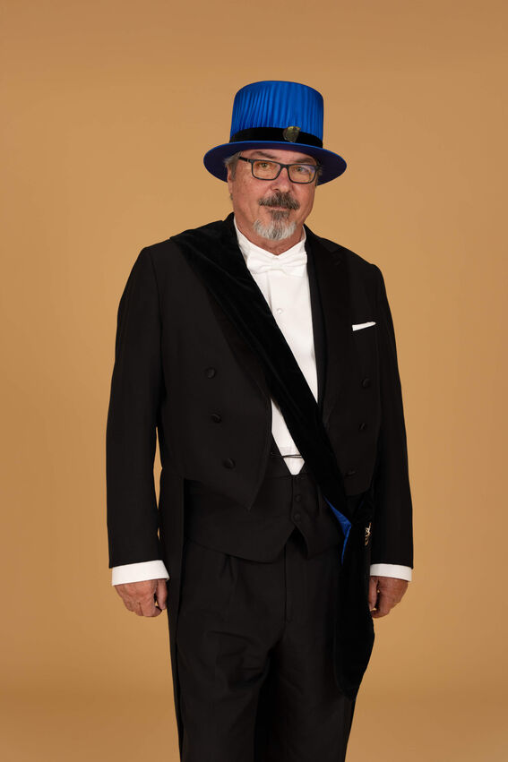 Henkilö pukeutuneena mustaan pukuun, valkoiseen paitaan ja siniseen hattuun, seisoo vaaleanruskean taustan edessä.